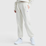 A person in Champion Powerblend Oversized Sweatpants with a C logo stands against a plain background, wearing off-white fleece pants with an elastic waistband, a matching sweatshirt, and white sneakers. One hand rests in their pocket.
