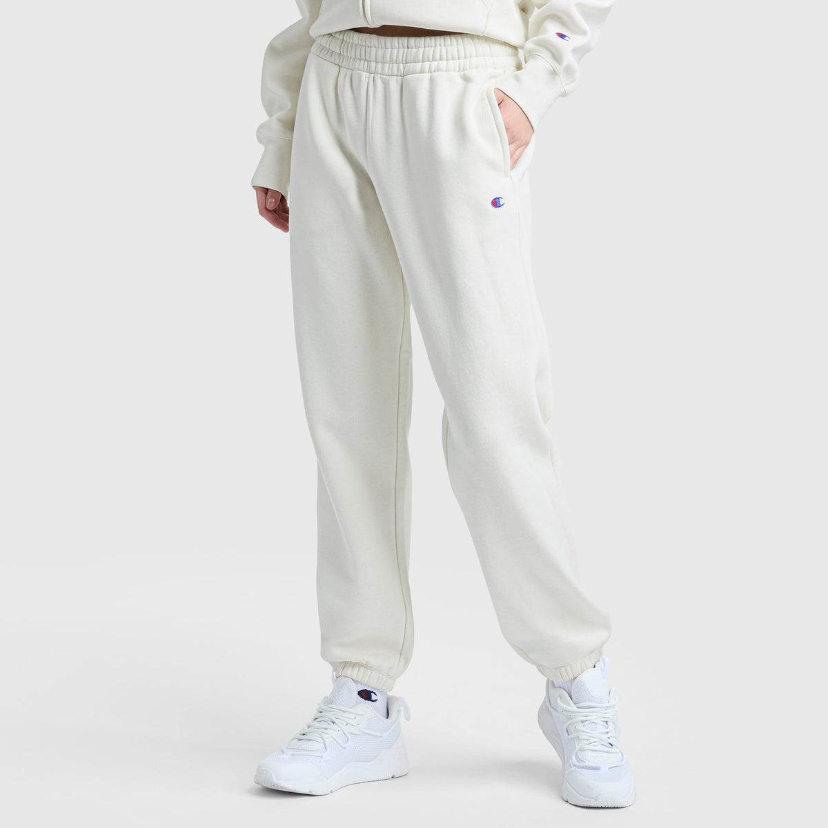 A person in Champion Powerblend Oversized Sweatpants with a C logo stands against a plain background, wearing off-white fleece pants with an elastic waistband, a matching sweatshirt, and white sneakers. One hand rests in their pocket.