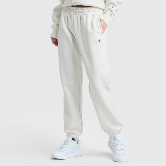 A person in Champion Powerblend Oversized Sweatpants with a C logo stands against a plain background, wearing off-white fleece pants with an elastic waistband, a matching sweatshirt, and white sneakers. One hand rests in their pocket.