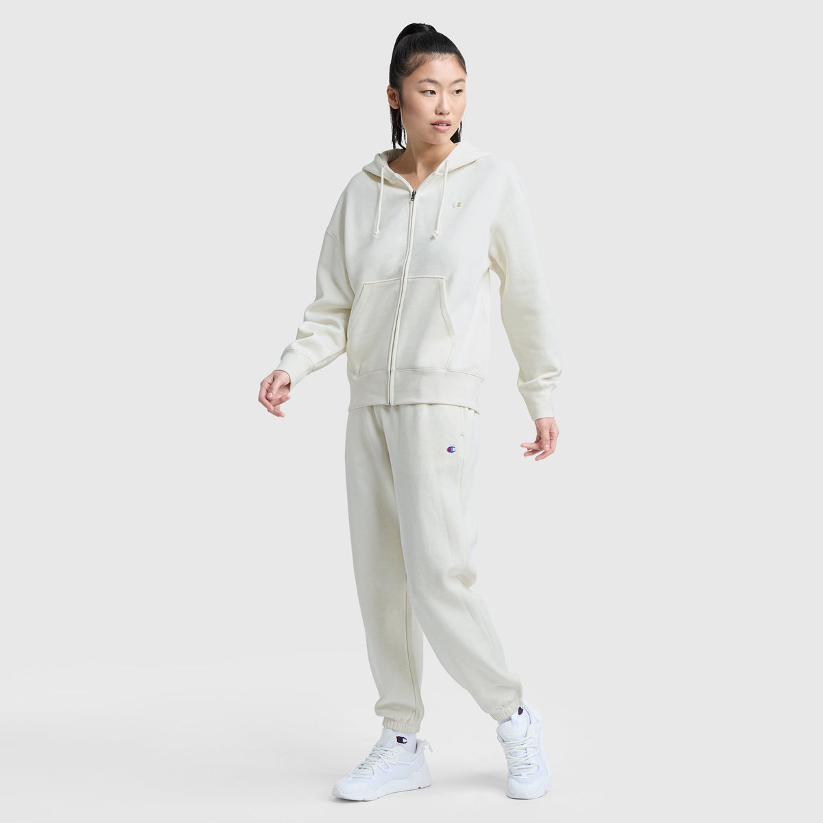 A person models Champion’s Powerblend Oversized Sweatpants with C Logo in off-white, paired with a matching zip-up hoodie and white sneakers, standing against a plain light background and gazing to the side.