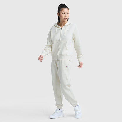 A person models Champion’s Powerblend Oversized Sweatpants with C Logo in off-white, paired with a matching zip-up hoodie and white sneakers, standing against a plain light background and gazing to the side.