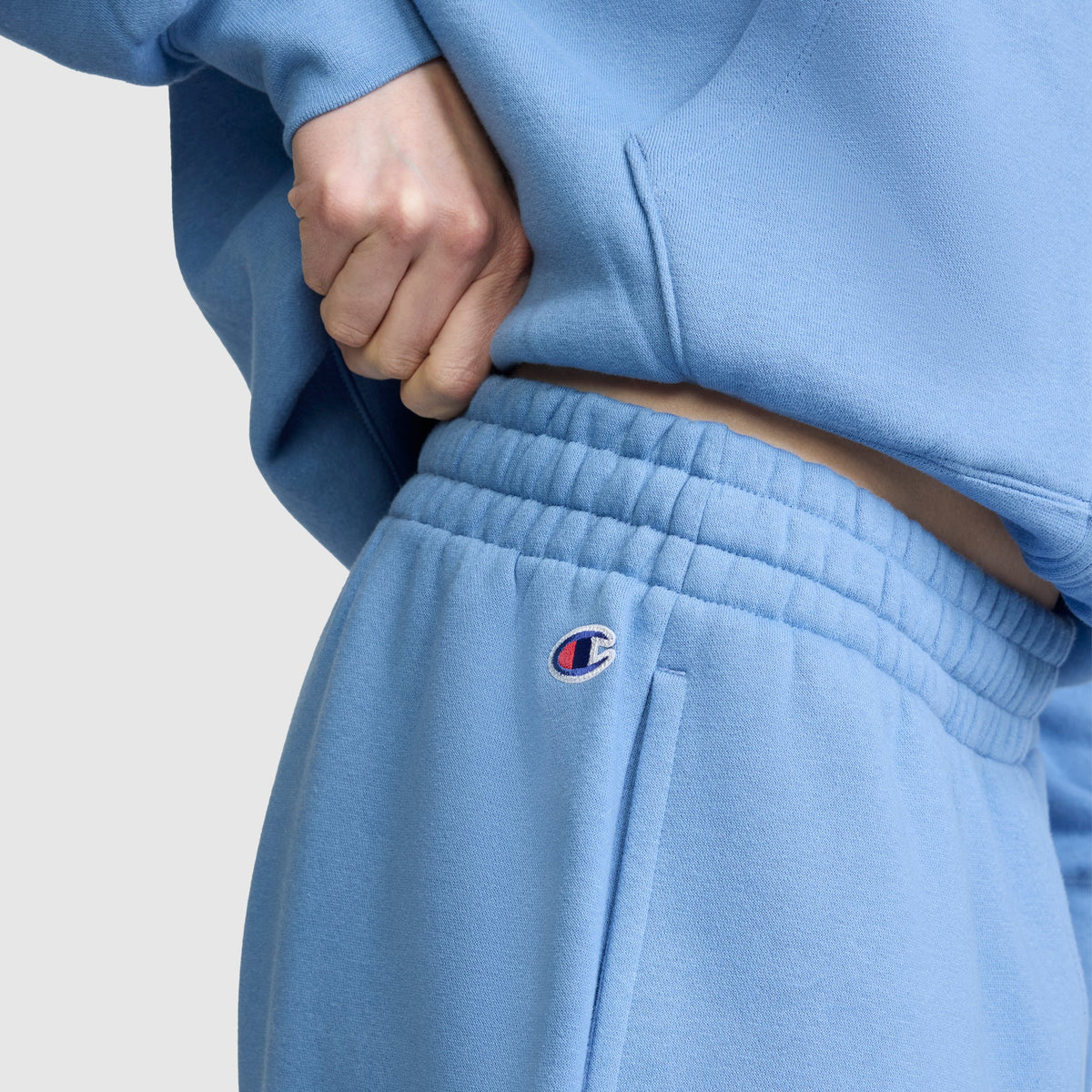 Wearing Champion’s Powerblend Oversized Sweatpants with the signature C logo in light blue, a person adjusts the waistband of the ultra-soft fleece pants. The cozy sweatpants feature an oversized fit and a small Champion logo on the leg.