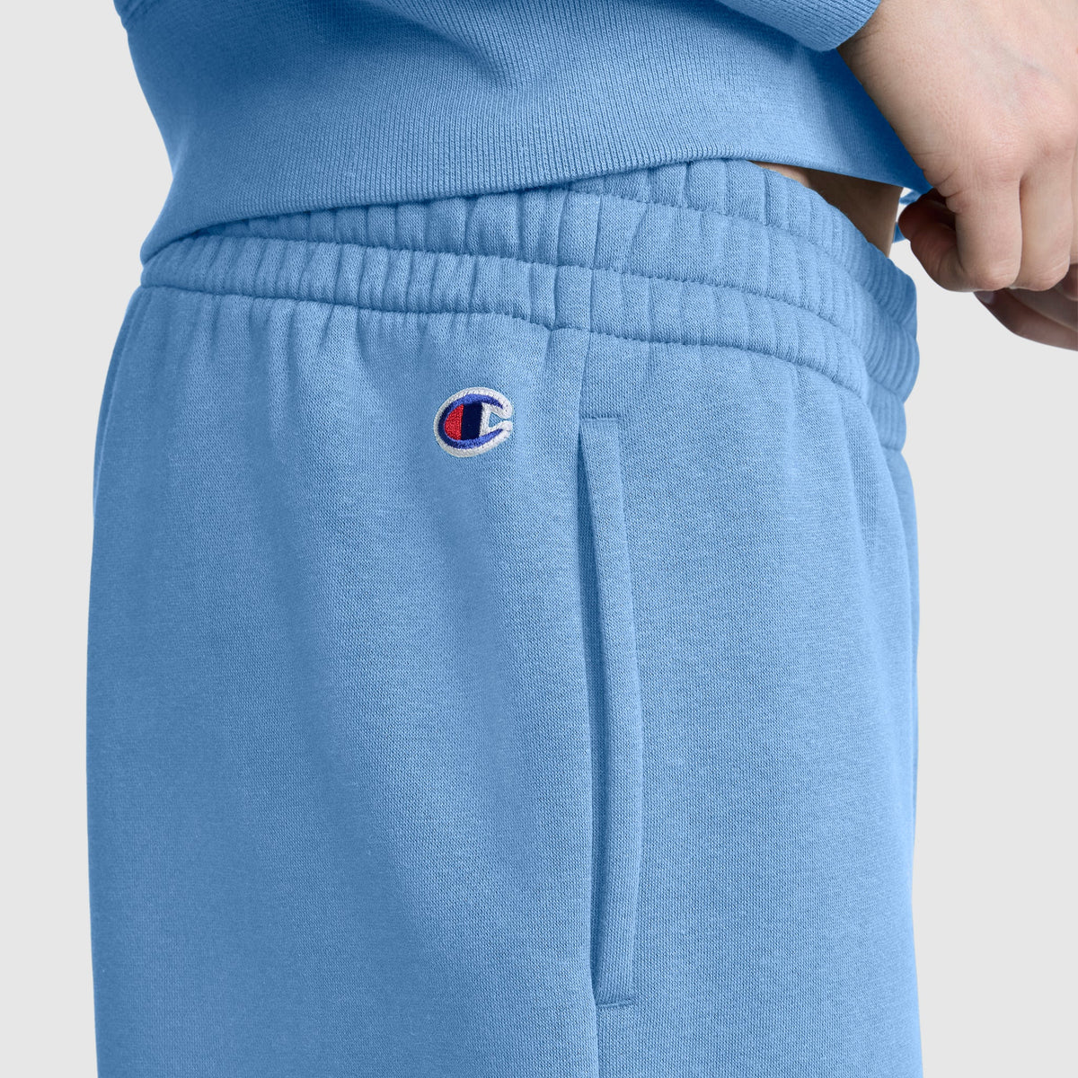 Wearing Champion’s Powerblend Oversized Sweatpants, C Logo in light blue, a person pairs the cozy fleece pants with a matching sweatshirt. The small Champion C logo is embroidered near the pocket, and their hand rests casually at the waistband, showcasing both comfort and style.