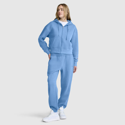 A person wearing a light blue hooded sweatshirt and Champion Powerblend Oversized Sweatpants with the C logo stands with one hand in their pocket. They also wear white sneakers and a white t-shirt, posing against a plain light gray background.