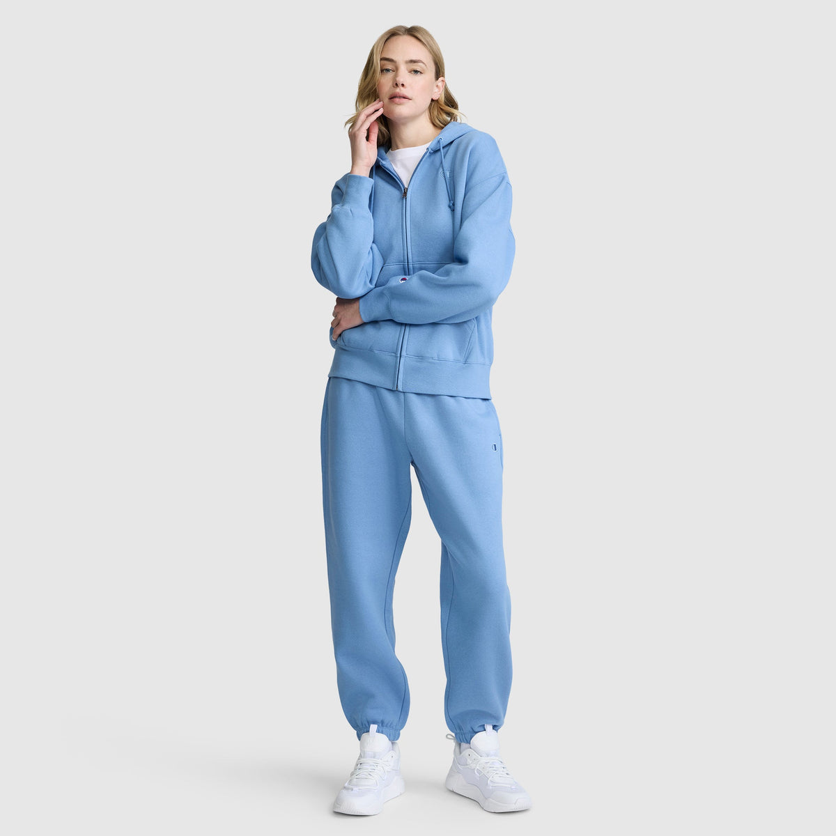 A woman stands against a plain light gray background, touching her face. She is wearing Champion’s Powerblend Oversized Sweatpants with the signature C Logo in light blue, paired with a matching hoodie and white sneakers.