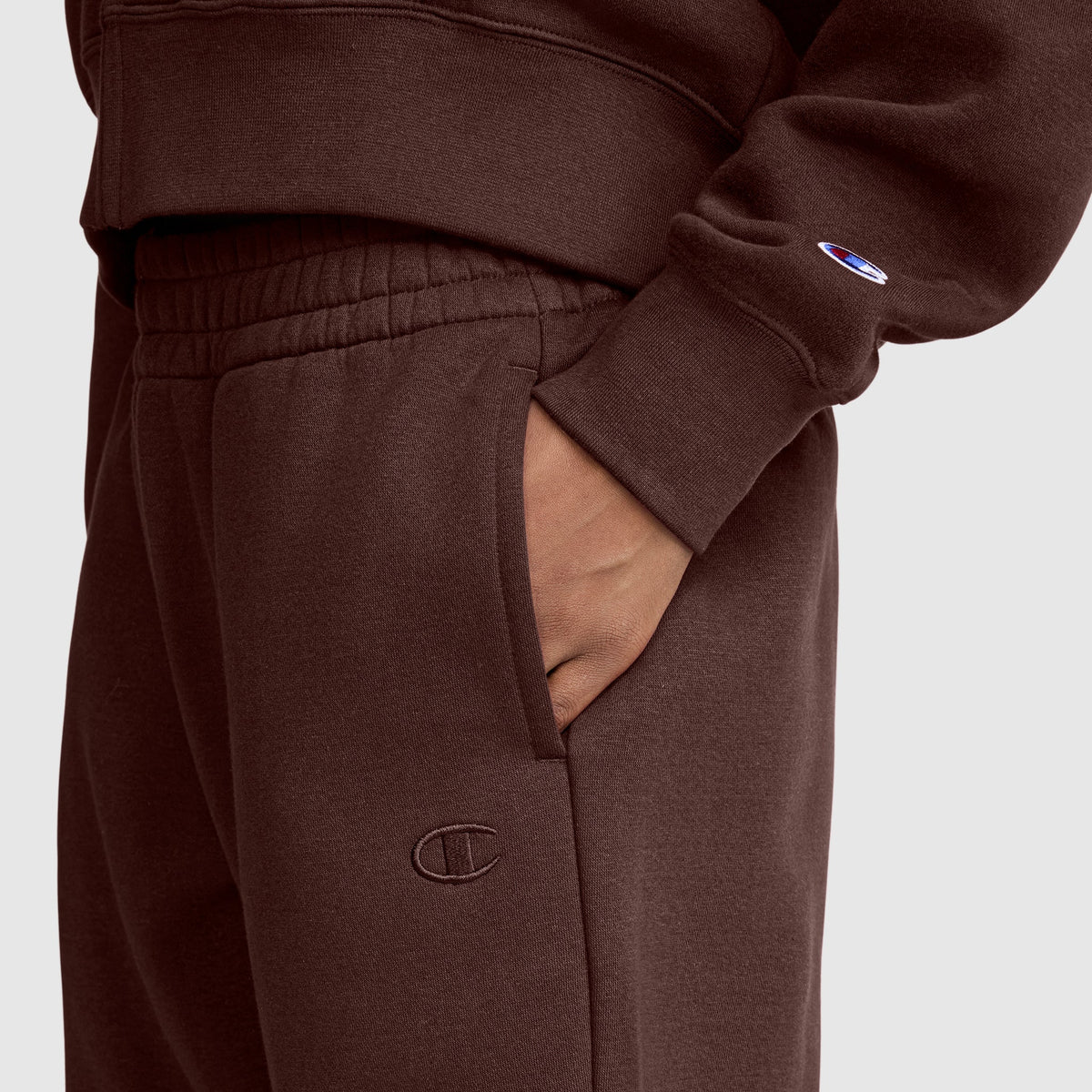 A person wears a brown Champion tracksuit. Their hand is in the pocket of the pants, which are the Champion Powerblend Oversized Sweatpants featuring the iconic C Logo. The sleeve displays a small embroidered Champion emblem. The oversized sweatpants give a relaxed look, set against a plain, light-colored background.