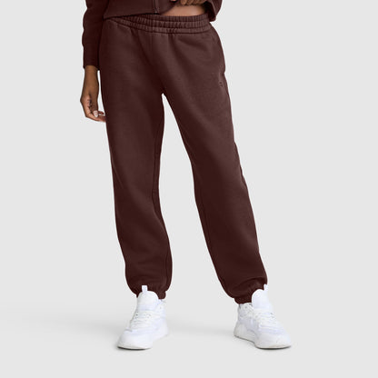 A person wearing maroon Champion Powerblend Oversized Sweatpants with a C Logo and white sneakers stands against a plain light gray background. Only the lower body and part of one arm are visible.