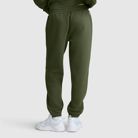 A person wearing Champion Powerblend Oversized Sweatpants with the C Logo in olive green and white sneakers stands facing away from the camera against a plain, light gray background.