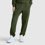 A person wearing Champion Powerblend Oversized Sweatpants, C Logo in olive green and white sneakers stands against a plain light gray background, with only the lower half of their body visible.