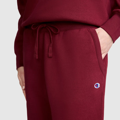 A person is wearing maroon Champion Powerblend Joggers, C Logo, paired with a matching sweatshirt and has one hand in their pocket. The Powerblend fleece joggers feature a small embroidered Champion C logo near the pocket. The background is plain and light-colored.