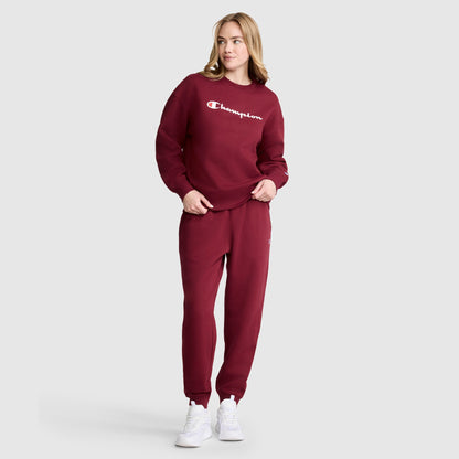 A woman wearing a maroon Champion sweatshirt and matching Powerblend Joggers, C Logo by Champion stands with one hand in her pocket, looking to the side against a plain light gray background.