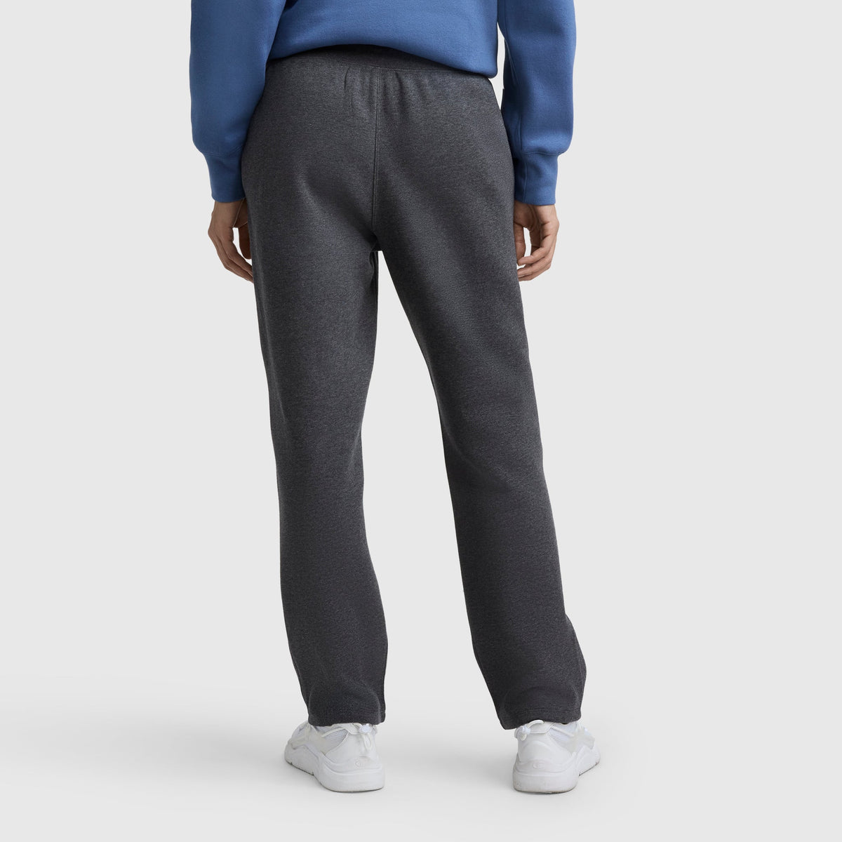 A person stands facing away in Champion Powerblend Straight Hem Sweatpants (dark gray), a blue sweatshirt, and white sneakers, set against a plain light background.