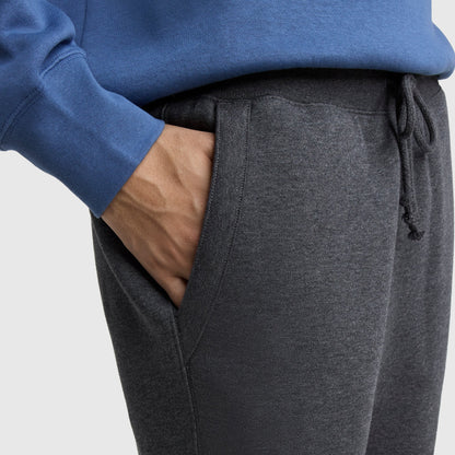 Someone is shown from the waist down against a plain background, wearing Champion Powerblend Straight Hem Sweatpants in dark gray with their right hand in their pocket and a blue sweatshirt.