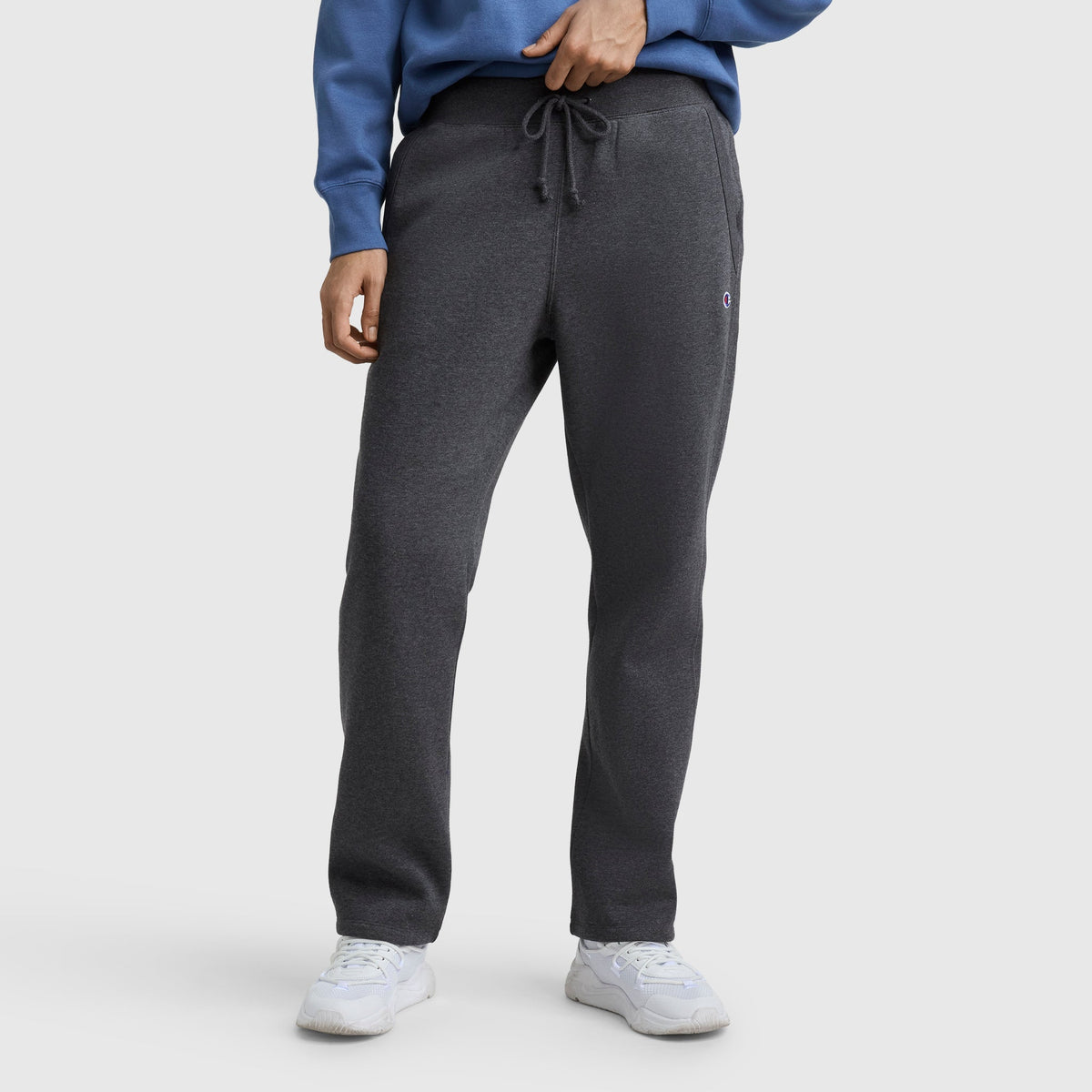 A person stands against a white background wearing a blue sweatshirt, white sneakers, and dark gray Champion Powerblend Straight Hem Sweatpants. The individual’s face is not visible.