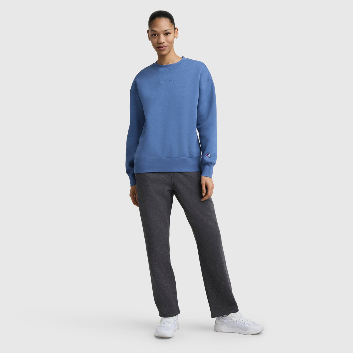 A person stands against a plain background, wearing a blue sweatshirt and Champion Powerblend Straight Hem Sweatpants in dark gray midweight fleece. White sneakers complete the look as they face the camera with a slight smile, hair tied back.