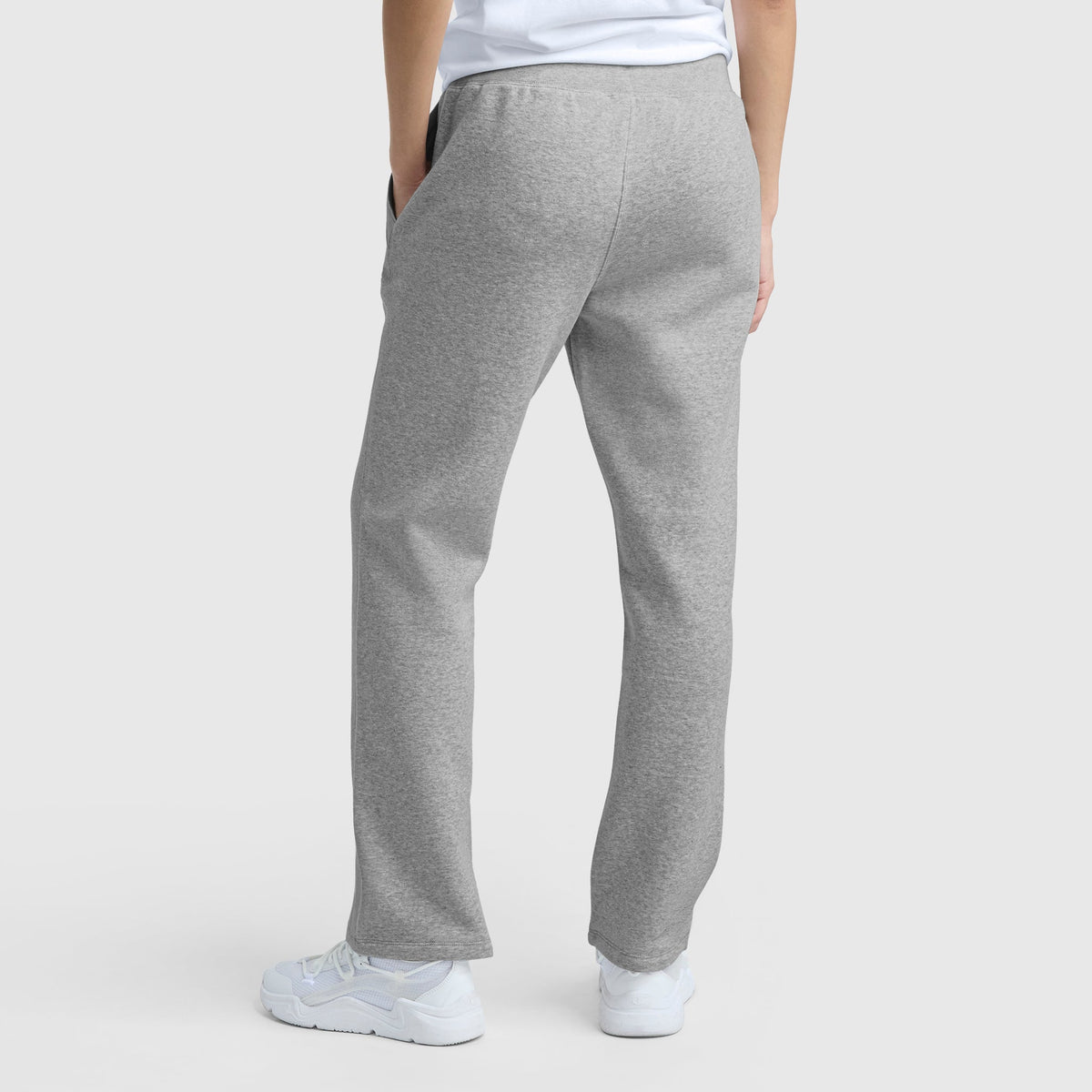 A person stands with their back to the camera in light gray Champion Powerblend Straight Hem Sweatpants, paired with white sneakers and a white t-shirt, against a plain light gray background, showcasing the pants’ fit from behind.