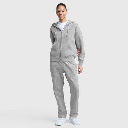 A person stands against a plain background wearing a light gray zip-up hoodie, Champion Powerblend Straight Hem Sweatpants, white sneakers, and a white t-shirt. They have a relaxed posture and a neutral expression.