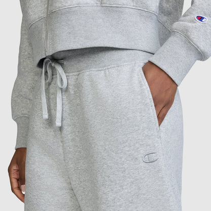 A person is wearing Champion’s Wide Leg Fleece Sweatpants featuring the signature C Logo, with their hand in the pocket; a matching sweatshirt from Champion is partially visible. Both pieces display the recognizable Champion branding. Set against a plain, light-colored background, these sweatpants offer a cozy and comfortable style statement.