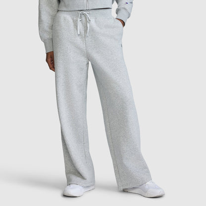 A person is wearing Champion's Wide Leg Fleece Sweatpants with the C Logo in light gray, paired with a matching sweatshirt and white sneakers. The photo is cropped from the shoulders down to showcase the relaxed fit and comfortable style of these sweatpants.