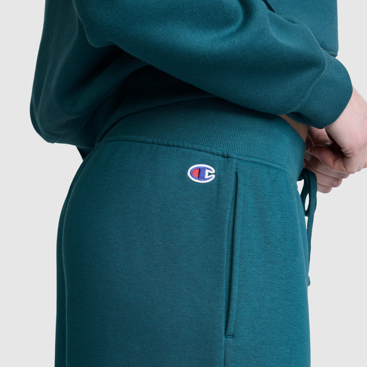 A person is wearing Champion’s Wide Leg Fleece Sweatpants, featuring the signature C logo near the pocket. The teal women’s sweatpants look cozy and are paired with a matching sweatshirt. The person’s hand rests near their hip against a plain light gray background.