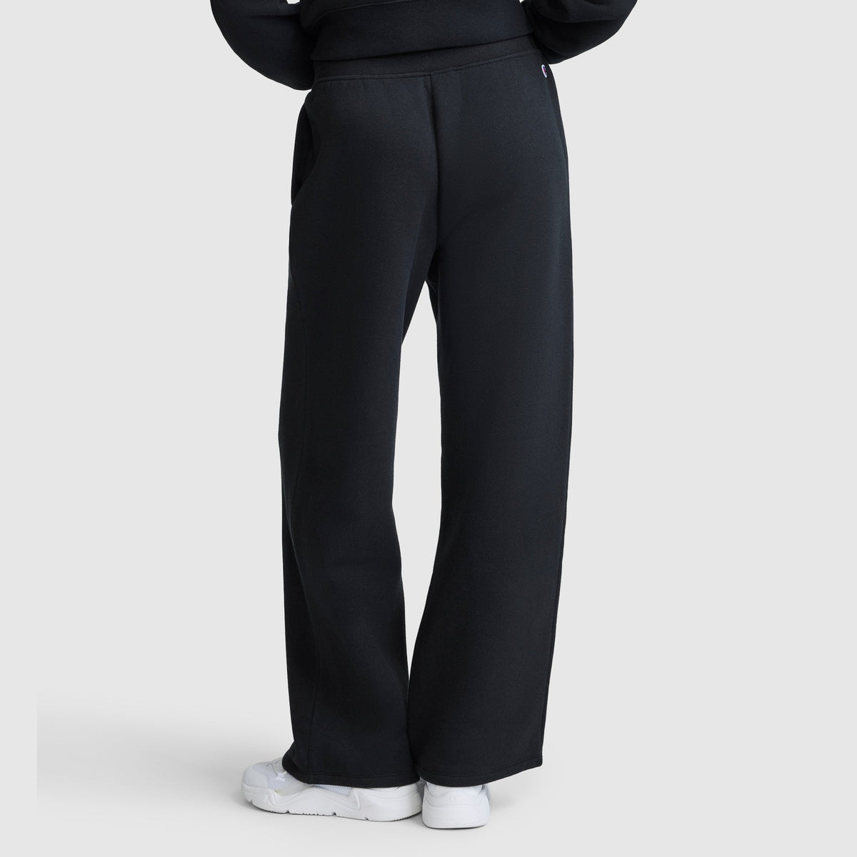 A person is seen from behind, standing against a light gray background, wearing Champion Wide Leg Fleece Sweatpants, C Logo in black paired with white sneakers. The full length of the women's sweatpants is visible in the photo.