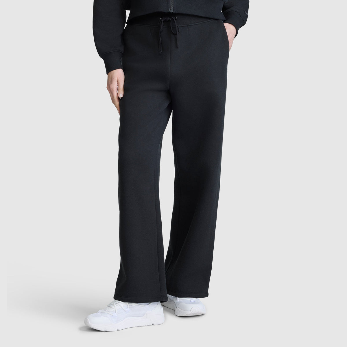A person wearing black Champion Wide Leg Fleece Sweatpants with a C Logo, a matching black sweatshirt, and white sneakers stands against a plain light gray background.