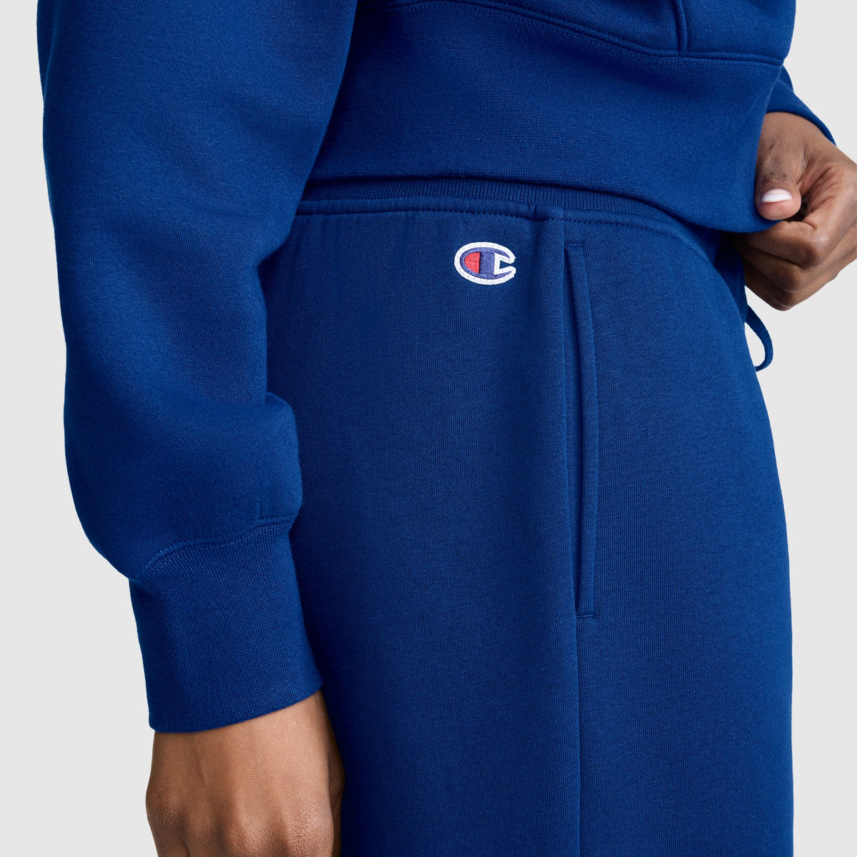 A person is wearing a blue Champion Wide Leg Fleece Sweatpants, C Logo, featuring the Champion logo above the pocket, standing against a plain light background.