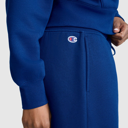 A person is wearing a blue Champion Wide Leg Fleece Sweatpants, C Logo, featuring the Champion logo above the pocket, standing against a plain light background.