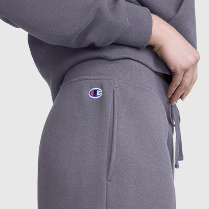 Close-up of a person wearing Champion’s Wide Leg Fleece Sweatpants, C Logo, in gray, paired with a matching sweatshirt. The pants feature a small Champion logo detail and the person has their hand resting on their hip.
