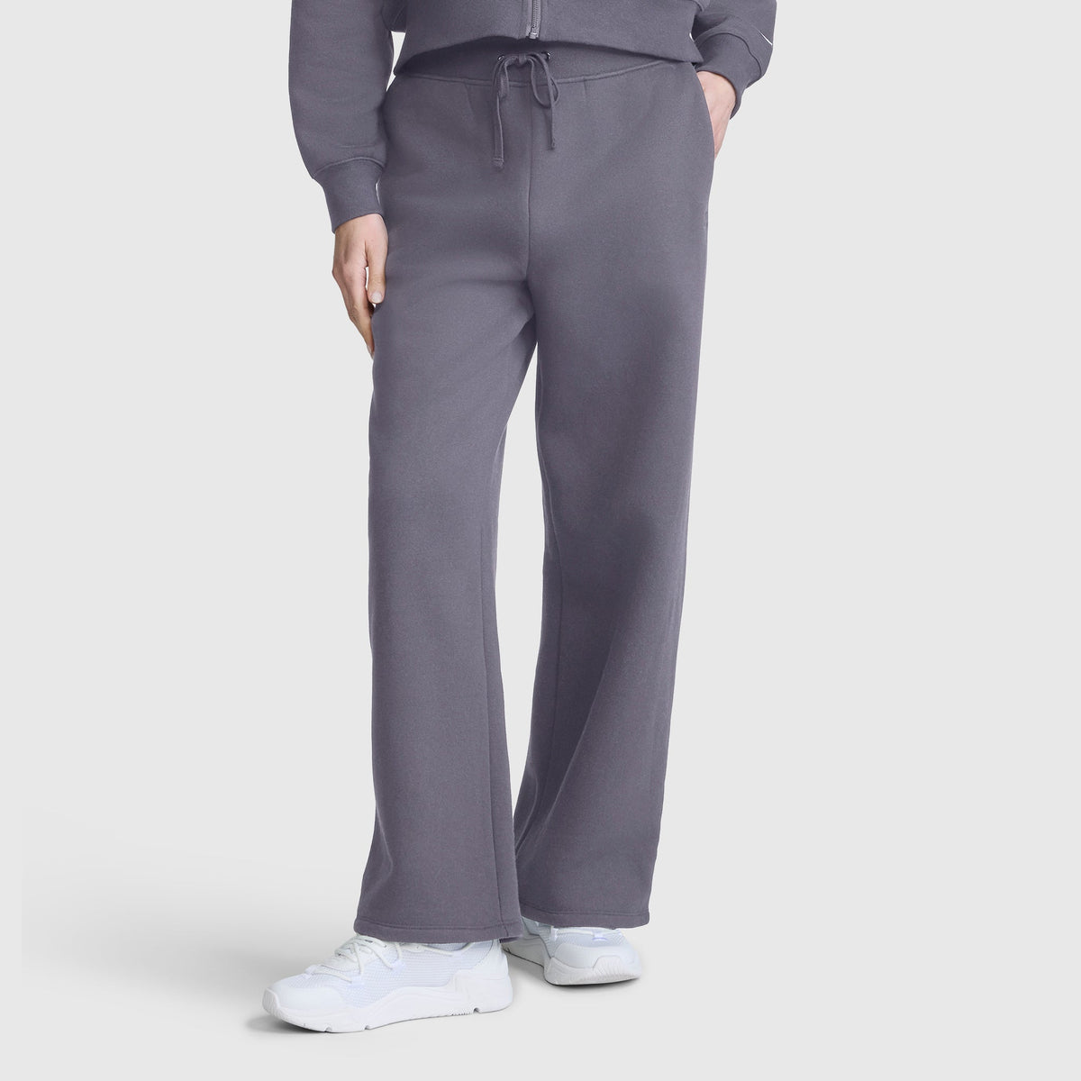 A person is shown from the waist down wearing loose-fitting Champion Wide Leg Fleece Sweatpants, C Logo, in gray, paired with white sneakers and a matching gray jacket. The modern silhouette of the Champion sweatpants stands out against a plain white background.