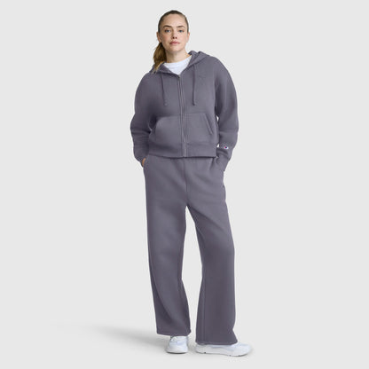 A person stands wearing Champion’s Wide Leg Fleece Sweatpants with the C Logo, styled as part of a matching gray set and paired with white sneakers. Their hands are in their pockets, and they gaze forward against a plain light background, highlighting the modern silhouette of the look.