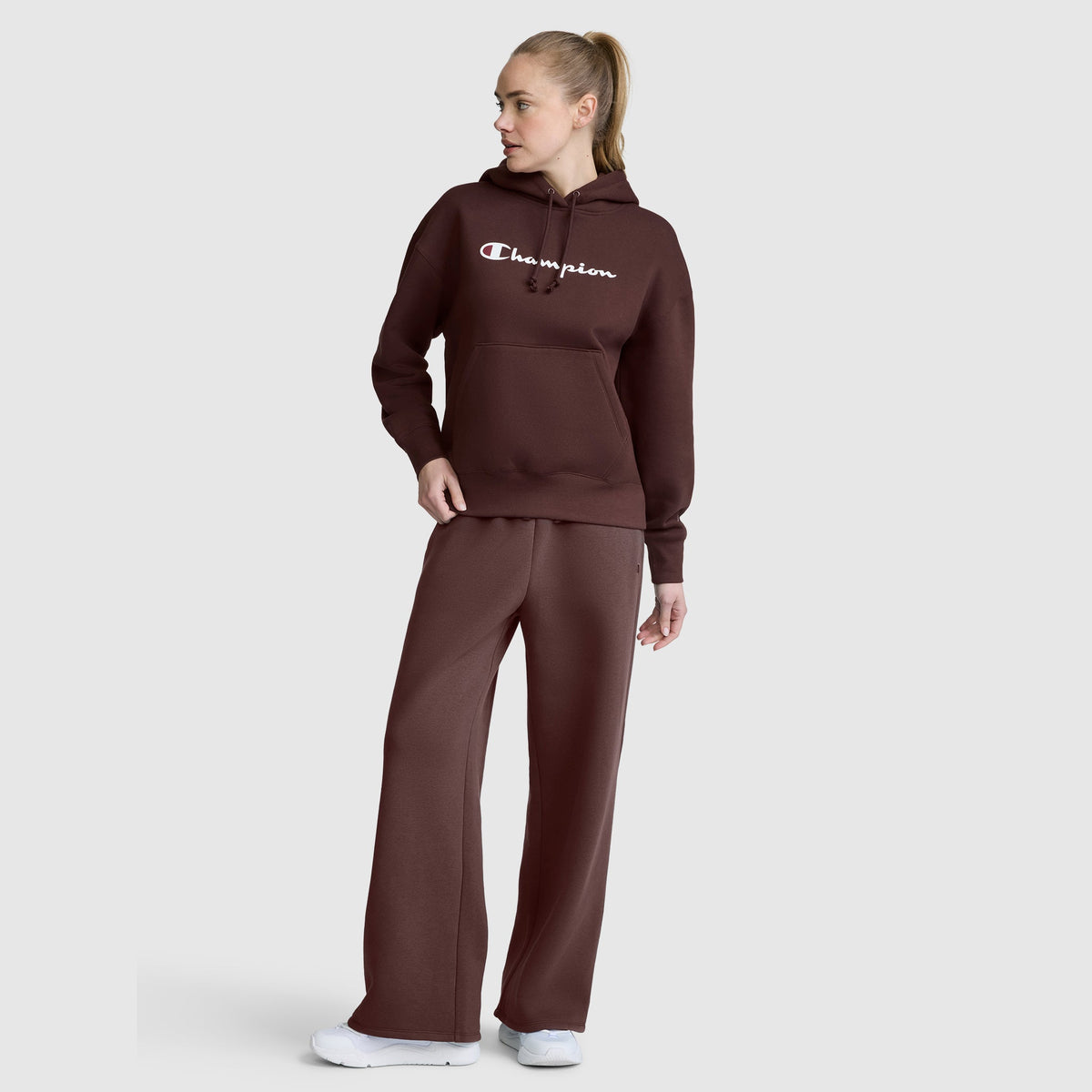 A woman with blonde hair in a ponytail stands sideways against a light gray background, looking off to her left. She is wearing white sneakers and a brown Champion hoodie paired with Champion Wide Leg Fleece Sweatpants, C Logo.