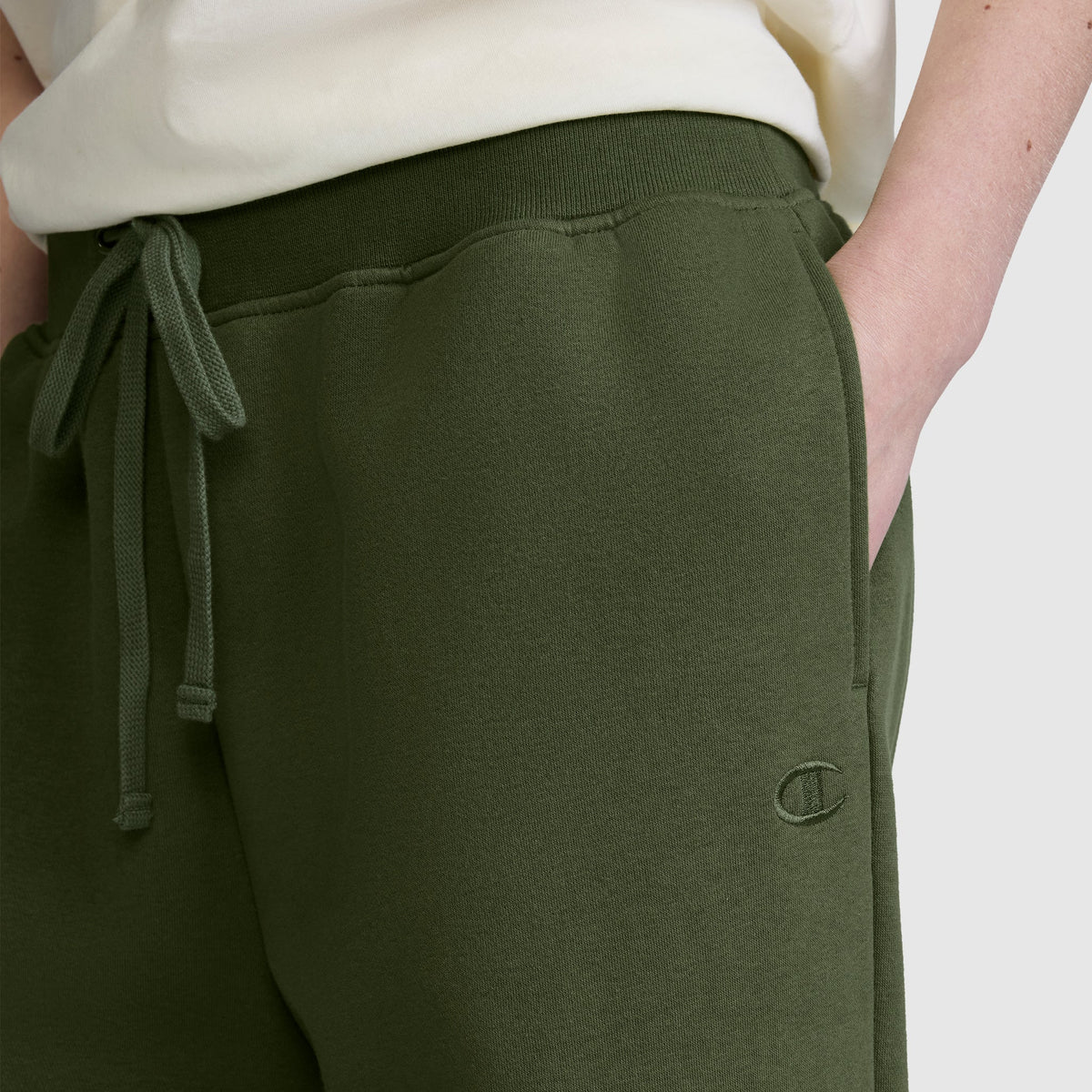 A person wearing Champion’s Wide Leg Fleece Sweatpants with the signature C Logo in olive green, featuring a drawstring waist and a relaxed wide leg fleece style; one hand in the pocket and a white shirt partially visible.