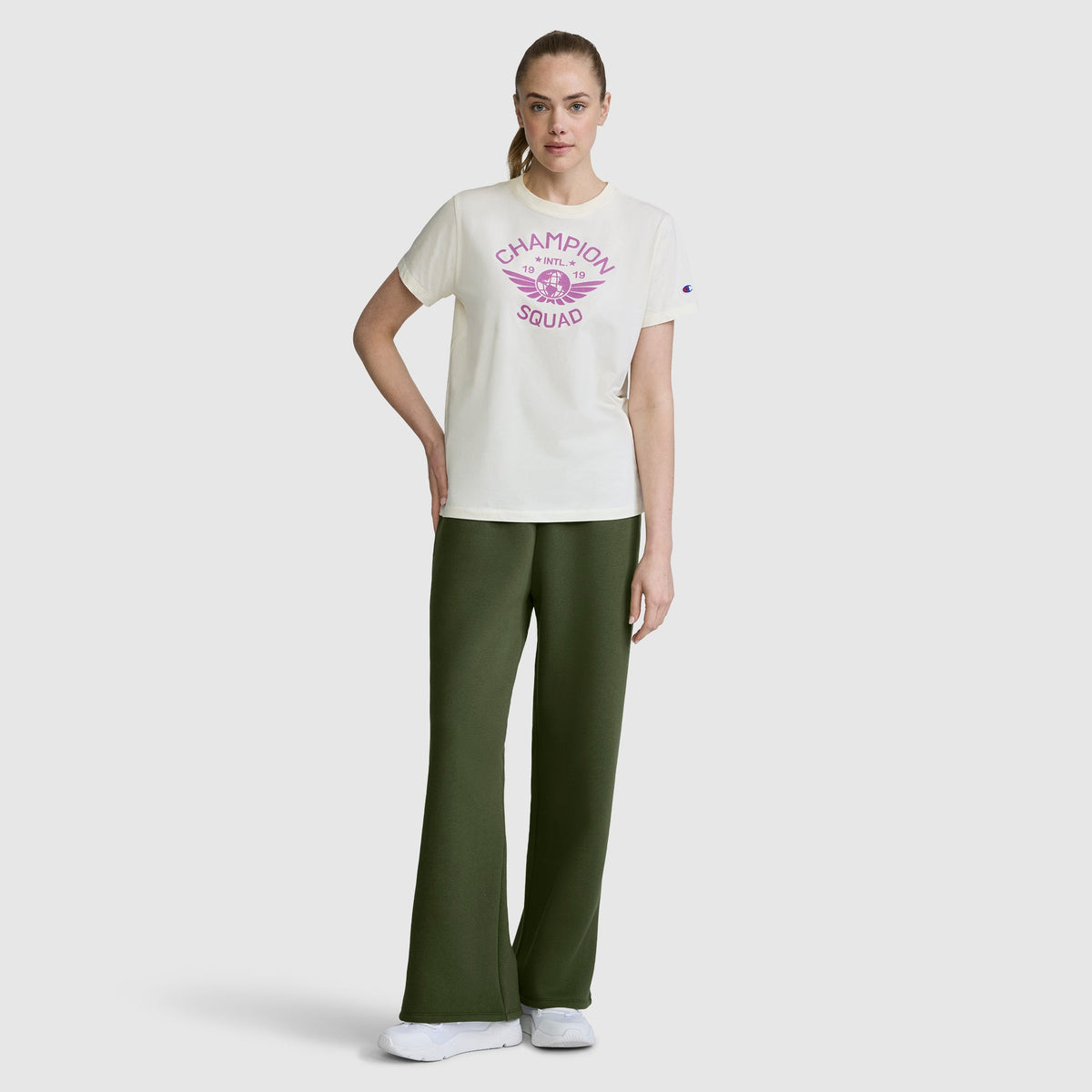 A woman stands against a plain light background, wearing a white Champion Squad t-shirt with pink lettering paired with Champion Wide Leg Fleece Sweatpants featuring the C Logo in dark green, and white sneakers. One hand rests on her hip while her other arm is relaxed.