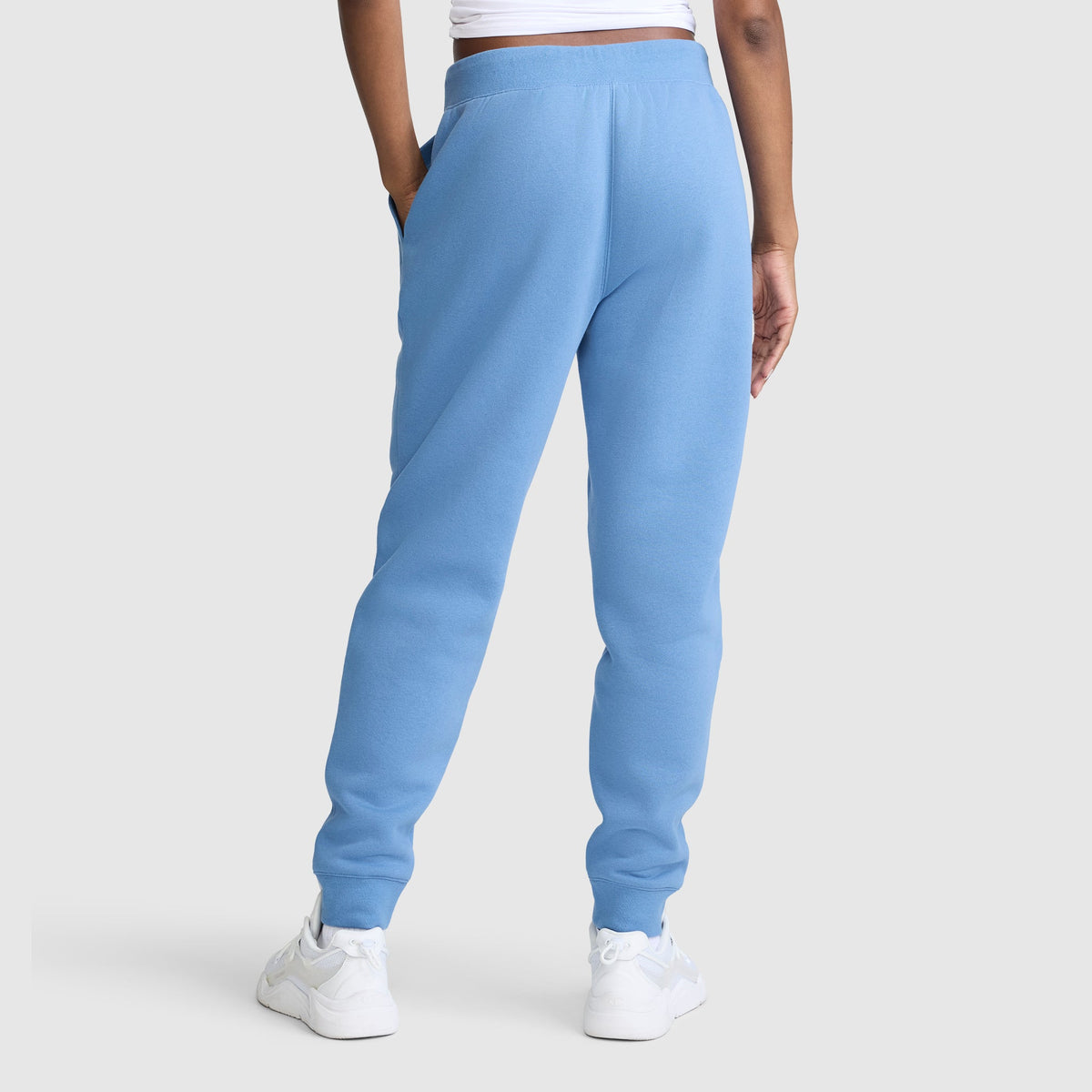 A person is shown from the back against a plain light gray background, wearing Champion Powerblend Joggers with the Classic Script Logo in light blue, paired with a white cropped top and white sneakers.