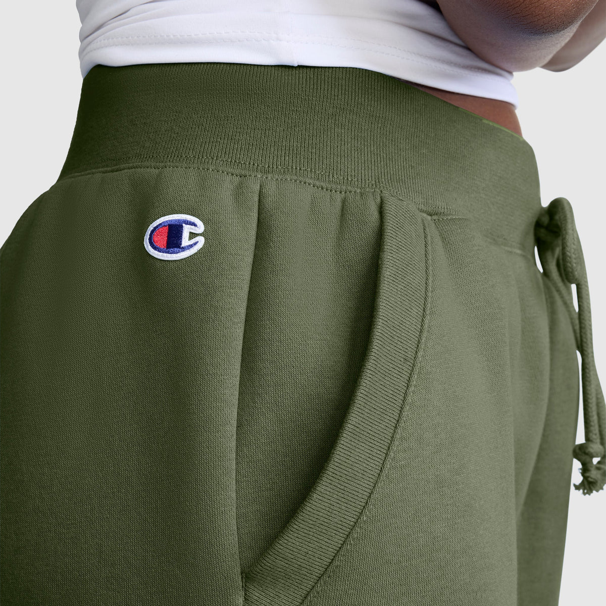 Close-up of a person wearing Champion Powerblend Joggers with the Classic Script Logo in olive green, featuring a white waistband and their hand resting in the pocket—highlighting these comfortable, everyday pants.