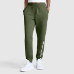 A person wearing Champion Powerblend Joggers for women in olive green, featuring the classic script logo in white on the left leg, paired with white sneakers and a white crop top. Their hands are in the joggers' pockets, set against a light gray background.