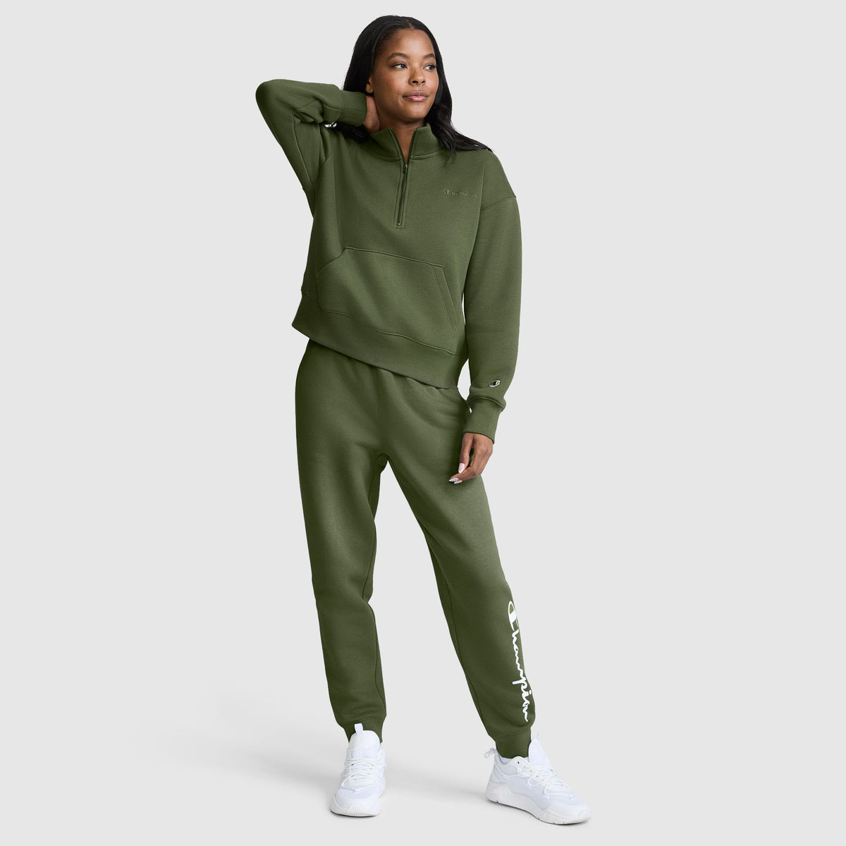 A woman in an olive green tracksuit with white sneakers stands with one hand behind her head and the other by her side, posing against a plain light gray background. Her comfortable Champion Powerblend Joggers, featuring the classic script logo, add both style and ease to her relaxed look.