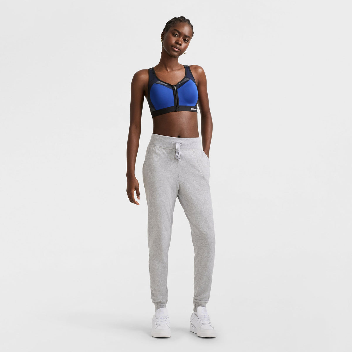 A woman stands against a plain white background wearing a blue sports bra, Champion Lightweight Lounge Joggers in light gray, and white sneakers, with her hands in her pockets.