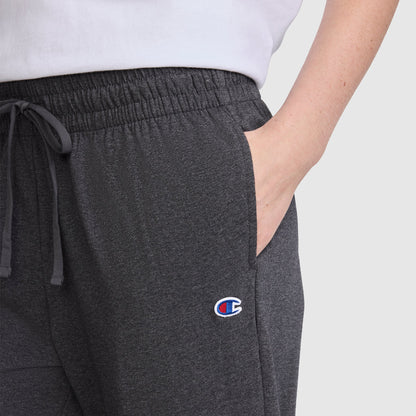 Wearing the Champion Lightweight Lounge Pants, C Logo in dark gray—renowned for their Champion quality and ultra-soft comfort—a person stands with one hand in the pocket. Only the lower half of a white shirt and a single arm are visible.