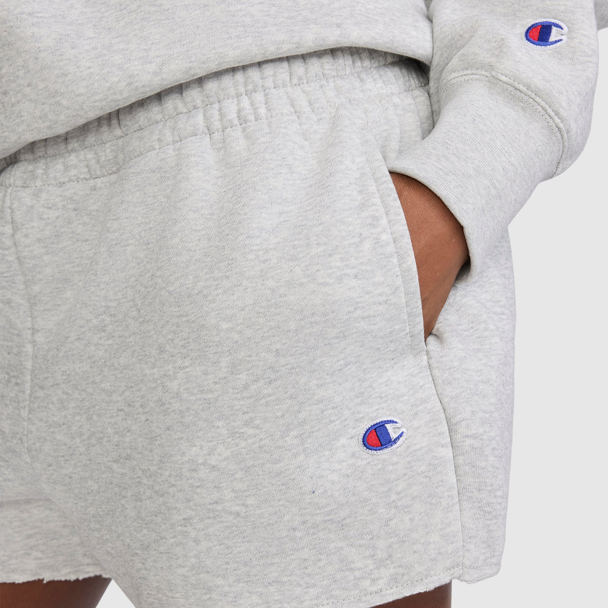 A person wears Champion's 3" Powerblend Shorts, C Logo in light gray along with a matching long sleeve sweatshirt. Both pieces display the signature blue and red Champion "C" logo for a classic touch. These comfortable Champion athletic shorts are ideal for everyday wear.