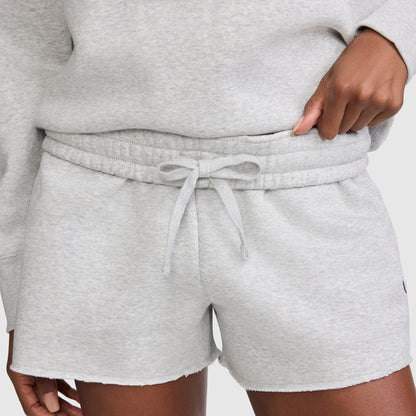 A person wearing Champion 3" Powerblend Shorts, featuring light gray fleece fabric with a drawstring waistband and the signature C Logo, paired with a matching sweatshirt. Only the lower torso and hand adjusting the waistband are visible.