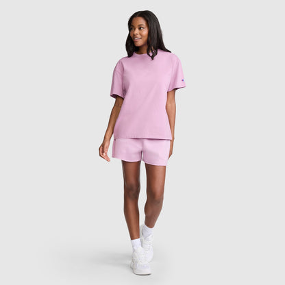 A woman wearing Champion’s 3" Powerblend Shorts, C Logo in light pink and a matching t-shirt smiles while walking. She has shoulder-length hair and completes her look with white socks and sneakers, standing against a plain light gray background.