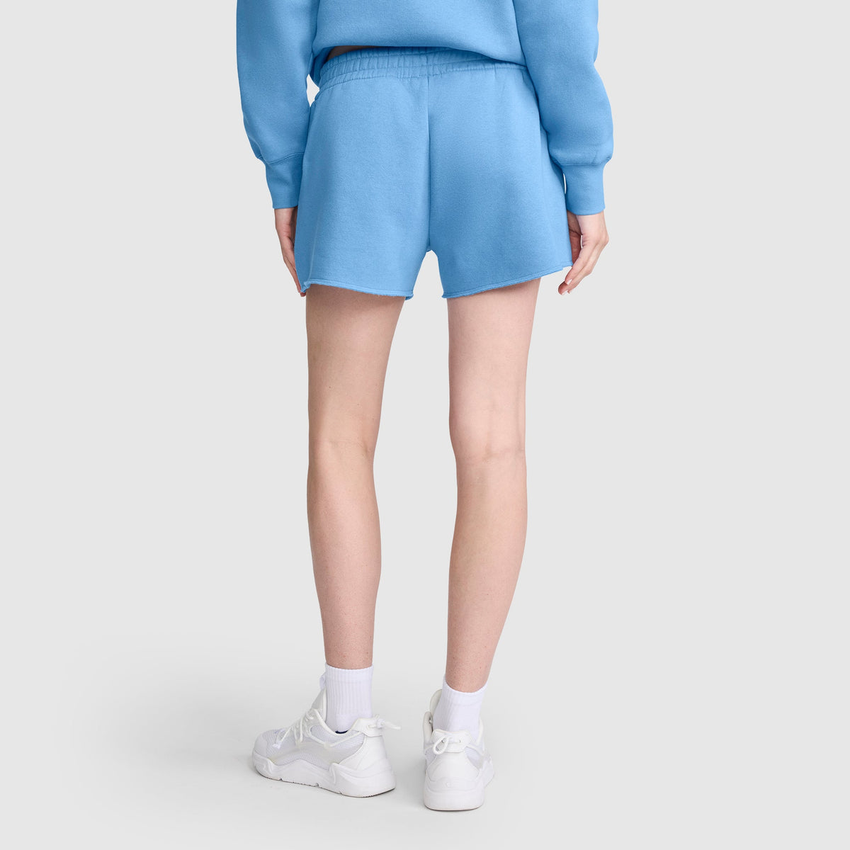 A person is standing facing away, wearing a blue sweatshirt and a pair of Champion 3" Powerblend Shorts with the C Logo, paired with white socks and white sneakers, set against a plain light background.