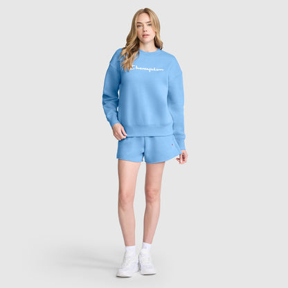 A person stands against a plain light gray background wearing a matching light blue sweatshirt and Champion 3" Powerblend Shorts featuring the signature C Logo, paired with white socks and white sneakers.