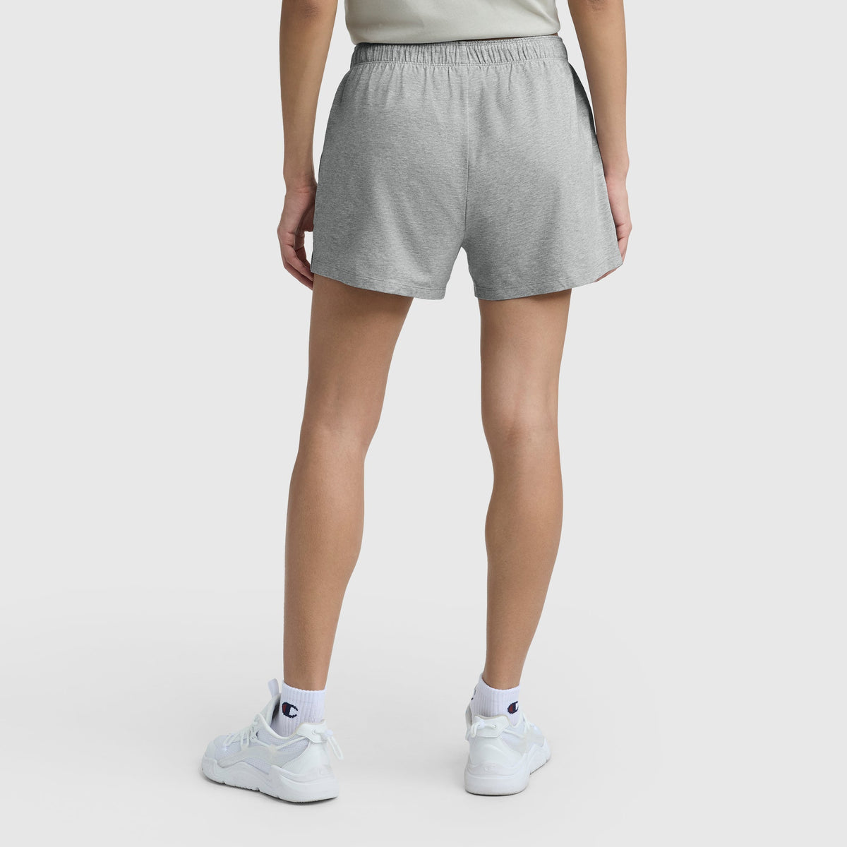 A person stands facing away, wearing Practice Shorts by Champion in light gray, with white sneakers and white logoed socks. The background is plain white.