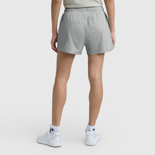 A person stands facing away, wearing Practice Shorts by Champion in light gray, with white sneakers and white logoed socks. The background is plain white.