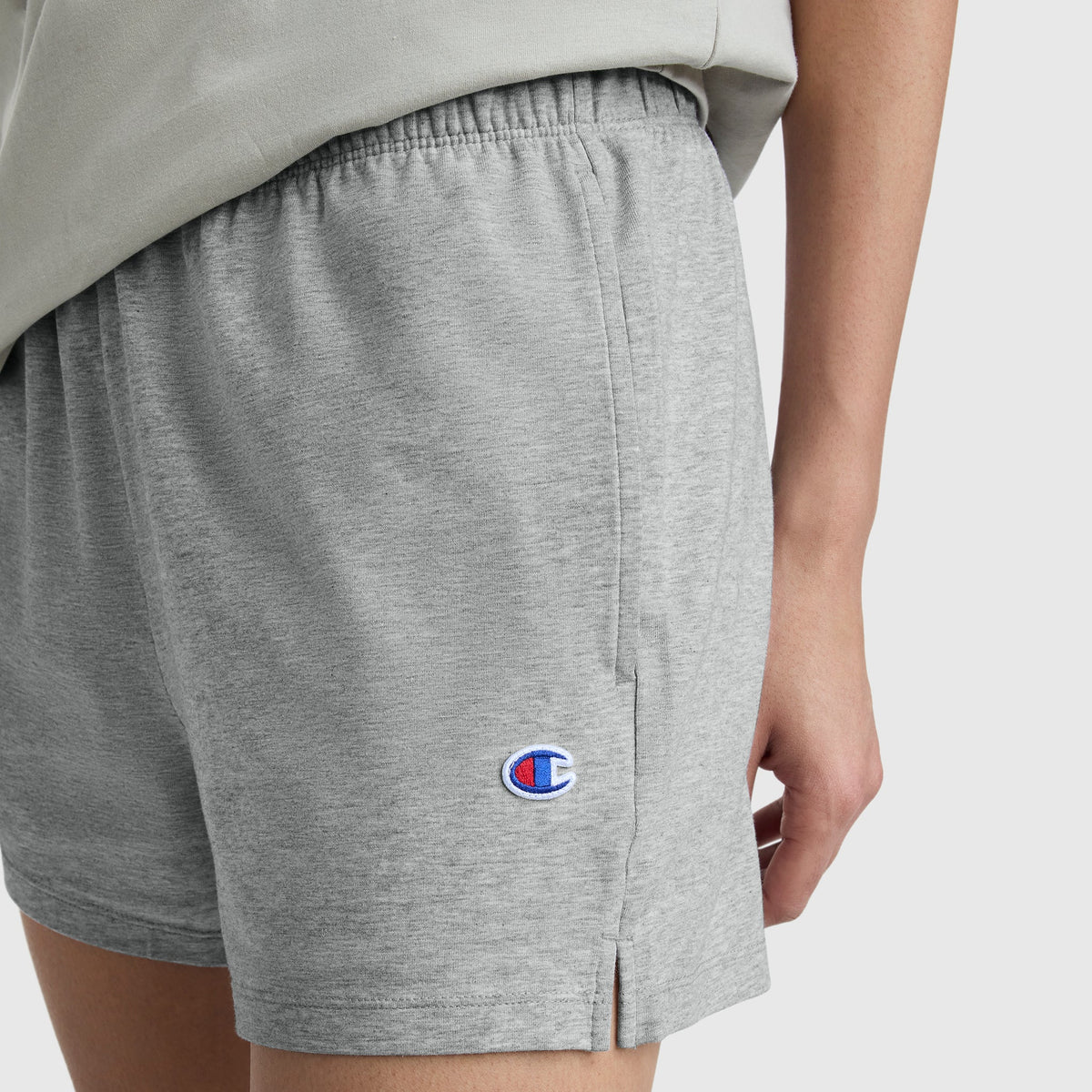 A person is shown in Champion Practice Shorts in light gray, with the logo near the hem, set against a plain background. Only their lower torso and part of one arm are visible.