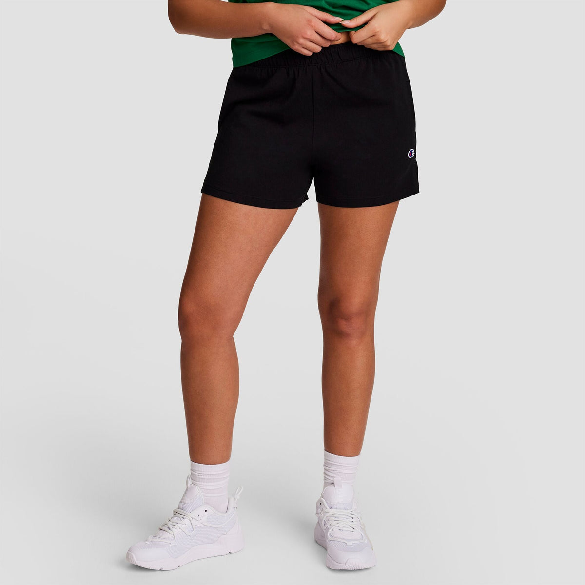 A person stands against a plain background, wearing a green shirt, Champion Practice Short in black, white socks, and white sneakers. The person's head is not visible in the image.