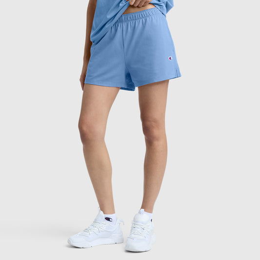 A person wearing Champion’s Practice Short in light blue, paired with a matching t-shirt and white athletic sneakers, stands against a plain light gray background.